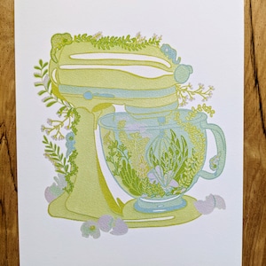 May include: A yellow stand mixer with a glass bowl filled with green and white flowers. The mixer is surrounded by green leaves and white flowers.