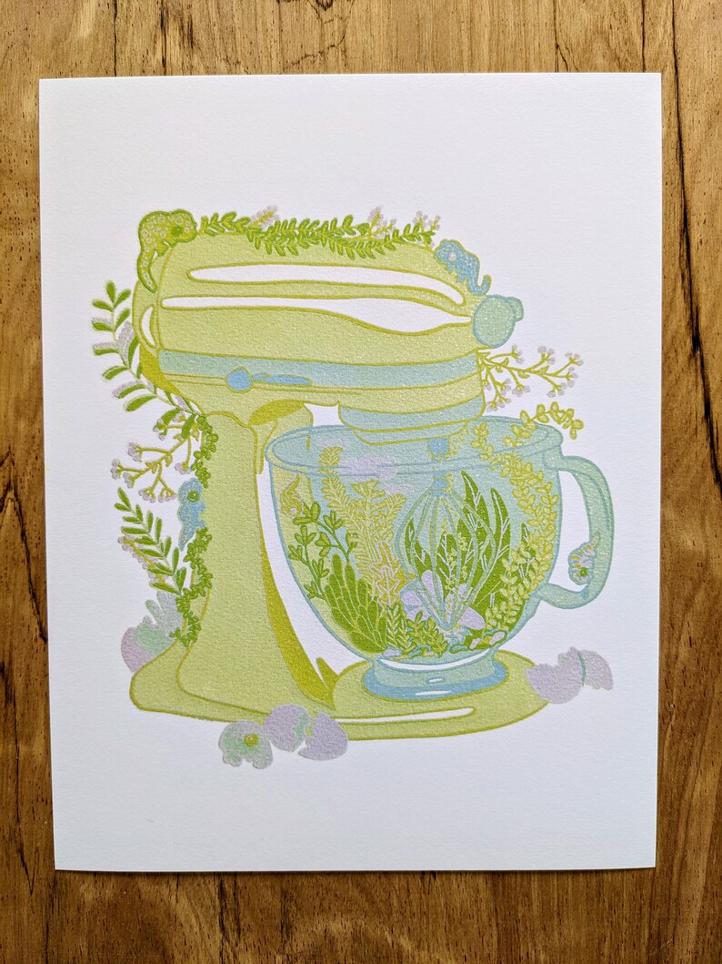 May include: A yellow stand mixer with a glass bowl filled with green and white flowers. The mixer is surrounded by green leaves and white flowers.
