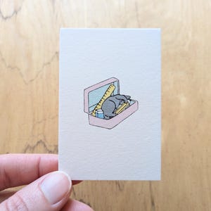 May include: A small rectangular card featuring a centered illustration of a pink pencil case containing a yellow ruler, a blue eraser, and a yellow pencil, with a small gray cat curled up inside the case among the school supplies.