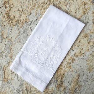 May include: A white rectangular linen napkin featuring a central embroidered wreath design in white thread. The wreath encircles a stylized letter S monogram. The fabric has a smooth texture with a visible hemmed edge along the bottom border.