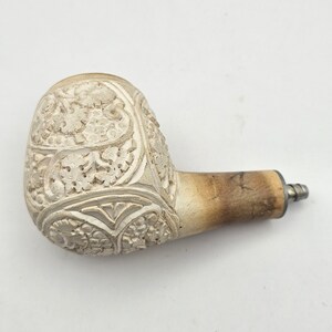 May include: A tobacco pipe featuring a cream-colored, carved bowl with intricate floral and leaf patterns. The stem transitions from a light, textured cream color to a smooth, darker tan finish, ending in a small, metallic silver-toned connector.