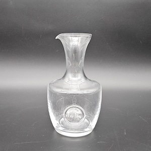 Puede incluir: Una decantador de vidrio transparente con cuerpo redondo y cuello corto. El decantador tiene un logotipo circular en la parte delantera que dice "Vino".