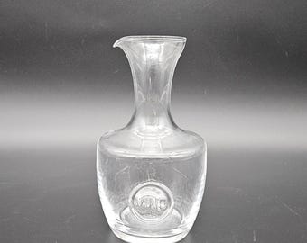 May include: A clear glass decanter with a round body and a short neck. The decanter has a circular logo on the front that says "Vino".