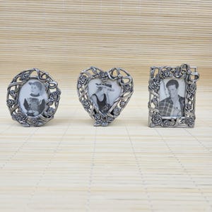 May include: Three silver-toned metal picture frames featuring intricate embossed rose and ribbon detailing. The set includes one circular frame, one heart-shaped frame, and one rectangular frame, each containing a black and white photograph.