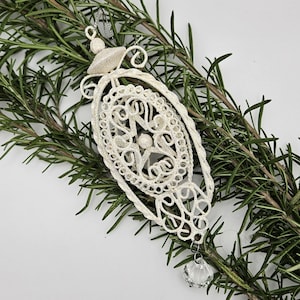 Könnte beinhalten: Weißer filigraner Weihnachtsbaumschmuck mit einem Kristalltropfen. Der Schmuck ist oval geformt und hat ein Sternmuster in der Mitte.