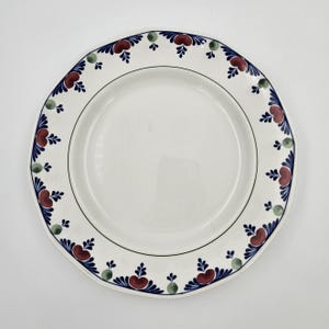 Może przedstawiać: Biały ceramiczny talerz z falistą krawędzią i ozdobnym obramowaniem. Obramowanie ma powtarzający się wzór czerwonych, niebieskich i zielonych motywów kwiatowych. Czarna linia podkreśla wewnętrzną krawędź talerza.
