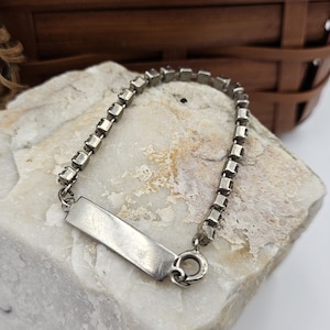 May include: A silver bracelet with a rectangular tag and a chain made of square-shaped rhinestones.