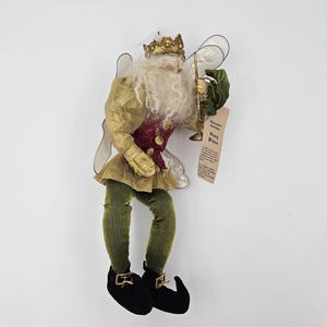 May include: A gold and green fairy prince ornament with a crown, white beard, and wings. He is holding a golden trumpet and has a tag that reads "Fairy Prince".