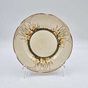 May include: Cream ceramic plate with a scalloped edge and gold trim. The plate features a central floral design of yellow daffodils and brown branches, set against a green border. A decorative piece for serving or display.