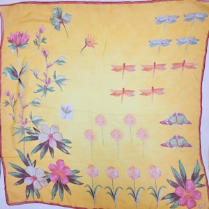 May include: A square yellow scarf featuring a hand-painted design of pink and white flowers with green leaves on the left and bottom edges. The right side displays rows of small orange and blue dragonflies and pink circular floral motifs.