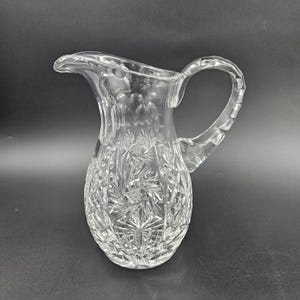 May include: A clear glass pitcher with a cut glass design. The pitcher has a round body and a handle.