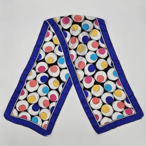 May include: A white silk scarf with a blue border and a pattern of colorful polka dots. The dots are various shades of pink, yellow, blue, and red.