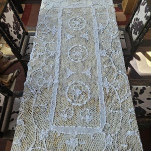 May include: Cream-coloured, rectangular lace tablecloth with intricate floral and geometric patterns. The lace features circular and star-shaped designs, and is laid out on a wooden table. Two chairs with cowhide-patterned seats are on either side.