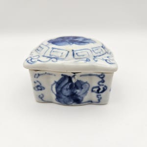 May include: A small, white ceramic box with a blue and white floral design. The box has a hinged lid and is decorated with a geometric pattern.