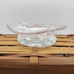 May include: A clear glass top hat-shaped container with a faceted base. The container is made of clear glass and sits on a woven bamboo surface. The top hat design adds a unique touch to the piece.
