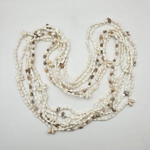 May include: A collection of multiple strands of white and tan seashell necklaces. The necklaces are made of small, natural seashells and are arranged in a layered pattern. The shells have a variety of shapes and sizes, with some featuring brown and tan markings.