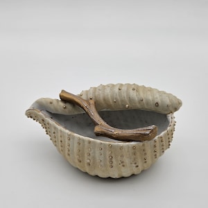 May include: A ceramic seashell-shaped dish with a gold-colored twig inside. The dish is decorated with small gold dots.