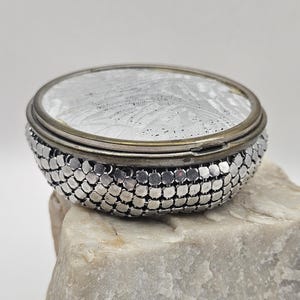 May include: A round metal compact case featuring a silver-toned mesh exterior with a textured, patterned lid. The lid has a metallic finish with a fine, intricate design, while the sides are constructed from small, interlocking silver-colored metal links.