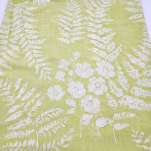 May include: A pale green fabric with a white floral and fern leaf design. The pattern showcases large, stylised white flowers and fern fronds against a soft, light green backdrop. The fabric seems to be a lightweight material, ideal for various textile projects.