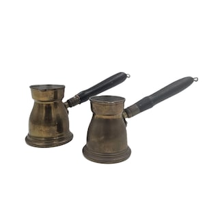 May include: Two antique brass coffee pots with black wooden handles. The pots have a classic, rounded shape and a spout for pouring. The handles are attached with metal rivets. The pots are displayed against a white background.