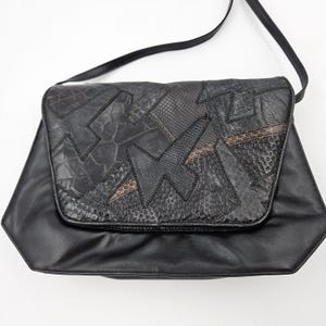 May include: Black leather shoulder bag with a geometric flap. The flap features a patchwork design with various textures and patterns, including faux snakeskin and crocodile. The bag has a long, thin strap and a structured shape.
