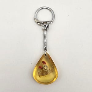 May include: A teardrop-shaped keychain pendant with a crab and small red objects encased in amber-colored resin. The pendant is attached to a silver-colored metal chain and keyring.