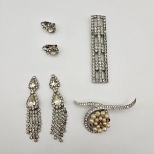 Könnte beinhalten: Eine Auswahl an Vintage-Schmuckstücken, darunter Clip-Ohrringe, Hängeohrringe und Broschen. Der Schmuck verfügt über klare Strasssteine und Kunstperlen, gefasst in silberfarbenem Metall. Die Stücke sind auf einer weißen Oberfläche angeordnet.