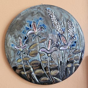 May include: A circular ceramic wall plaque featuring a relief design of stylized flowers and tall, textured stalks. The glaze is a mottled mix of grey, blue, and brown tones, with raised details highlighting the petals and leaves across the surface.