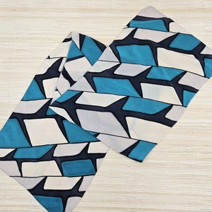 Op de afbeelding: Een rechthoekige stoffen sjaal met een herhalend geometrisch patroon van in elkaar grijpende vormen in tinten blauwgroen, gebroken wit en donker marineblauw. Het ontwerp bestaat uit gestileerde, hoekige vormen in een horizontale lay-out.