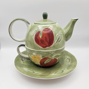 Op de afbeelding: Een groene keramische theepot en theekop set met een geschilderd appelontwerp. De theepot heeft een deksel en een handvat. De theekop heeft een handvat en staat op een bijpassend schoteltje.