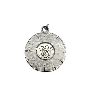 Puede incluir: Colgante plateado con un diseño circular texturizado. El colgante presenta una incrustación central circular con un monograma adornado. El borde exterior tiene un patrón texturizado en forma de estrella. Un pequeño lazo está en la parte superior para una cadena.