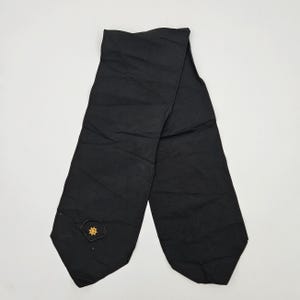 May include: A black necktie featuring a small, embroidered gold-colored ship's wheel emblem positioned near the pointed end. The tie has a smooth, matte fabric finish and a simple, clean design with a standard tapered shape and stitched edges.