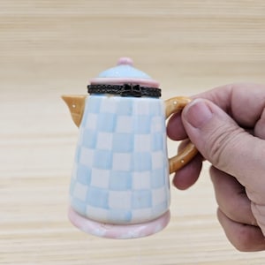 May include: A small ceramic teapot-shaped trinket box featuring a light blue and white checkered pattern. The lid is light blue with a pink rim and a dark metal hinge with a bow detail. The handle is tan with white dots, and the base is pink.