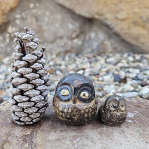 May include: Two ceramic owls, one larger than the other, with black and brown speckled paint and yellow eyes. The owls are sitting on a rock next to a pine cone.