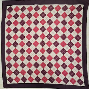 May include: A square scarf featuring an argyle pattern with alternating dark purple and bright pink diamonds on a cream background. The design includes thin, intersecting diagonal lines in a lighter shade, all framed by a solid dark purple border.