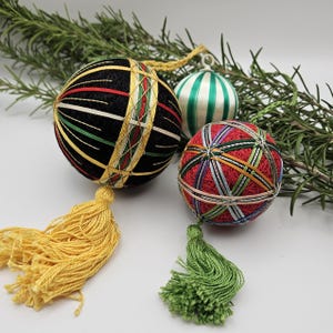 May include: Three handmade Christmas ornaments with colorful thread designs. The ornaments are round and have tassels. One ornament is black with green and gold thread, one is red with green and gold thread, and one is white with green thread.
