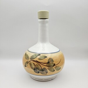May include: A white ceramic bottle with a tan and brown painted design of leaves and branches. The bottle has a round base and a long neck, topped with a beige cork. A blue stripe accents the design.