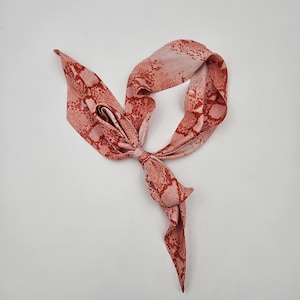 Op de afbeelding: Een roze en rode sjaal met slangenpatroon, in een strik geknoopt. De sjaal is gemaakt van een zacht, zijdeachtig materiaal en wordt gepresenteerd tegen een witte achtergrond. Het ontwerp van de sjaal heeft een herhalend patroon van rode schubben op een roze achtergrond.