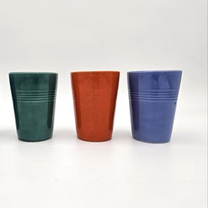 Op de afbeelding: Drie keramische bekers in groen, oranje en blauw. Elke beker heeft een cilindrische vorm met horizontale groeven aan de bovenkant. De bekers worden gepresenteerd tegen een witte achtergrond.