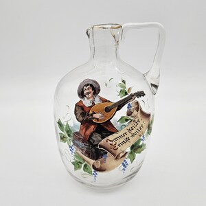 May include: A clear glass bottle with a handle, featuring a hand-painted scene of a man playing a lute. The bottle has a scroll with the text "Immer heiter, Trinke weiter!"  The bottle is decorated with green vines and blue grapes.
