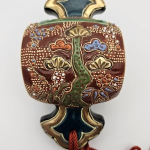 May include: A close-up of a decorative Japanese ornament with a red, green, and gold design. The ornament is made of ceramic and has a floral pattern. It is attached to a red cord.