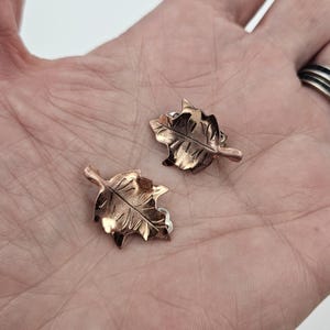 May include: A pair of copper-colored leaf-shaped earrings. Each earring features detailed veining and a textured surface, with a post backing. The earrings are displayed on a neutral background.
