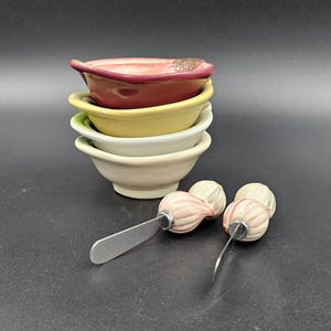 Può includere: Set di quattro ciotole in ceramica in tonalità di rosso, giallo e bianco, impilate l'una sull'altra. Sono inclusi due coltelli da burro in ceramica con manici a forma di cipolla.