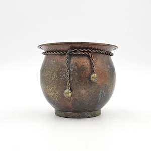 Peut inclure: Un petit pot rond en cuivre avec une patine sombre et usée. Le récipient présente une bordure décorative en cordelette de cuivre torsadée entourant le rebord supérieur, avec deux extrémités pendantes terminées par de petites perles métalliques.