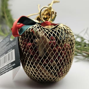 May include: A gold metal apple-shaped ornament with a red and green ribbon. The ornament is filled with small red berries and wood pieces.