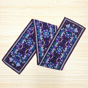 May include: A long, rectangular scarf featuring a deep burgundy background with a repeating pattern of white, purple, and teal floral and scrollwork motifs. The scarf has a dark border and a symmetrical design that runs along its entire length.