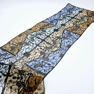 May include: A long, rectangular scarf with a repeating geometric pattern in shades of brown, light blue, and black. The scarf's design features ornate, scroll-like shapes. The scarf is made of a lightweight, sheer fabric.