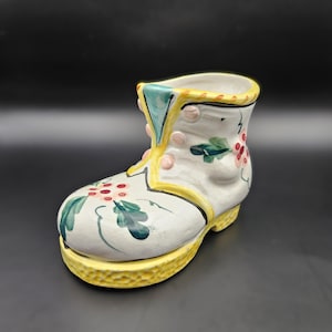 May include: A white ceramic boot planter with a yellow rim and floral designs in shades of green, red, and pink.