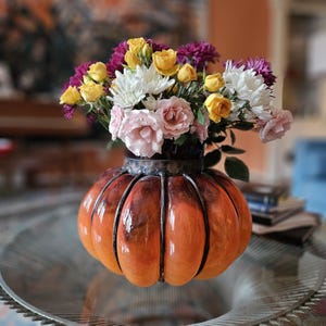 May include: A bouquet of yellow, white, and purple flowers in a brown and orange pumpkin-shaped vase.