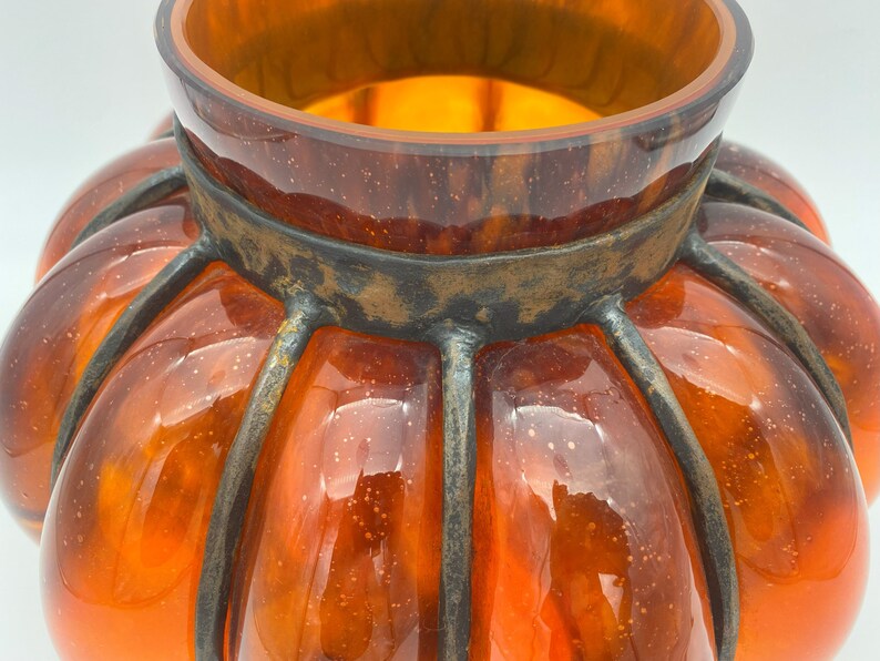 May include: Orange glass pumpkin-shaped vase with a metal cage around it. The vase has a textured surface and a shiny finish.